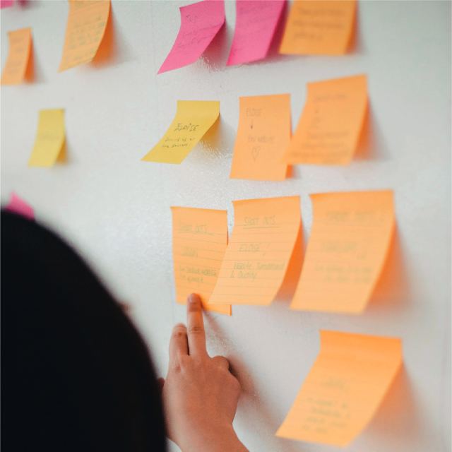 Pared blanca con trece post-it pegados, de color naranjo, amarillo y fucsia. Y una mano apuntando de uno de ellos